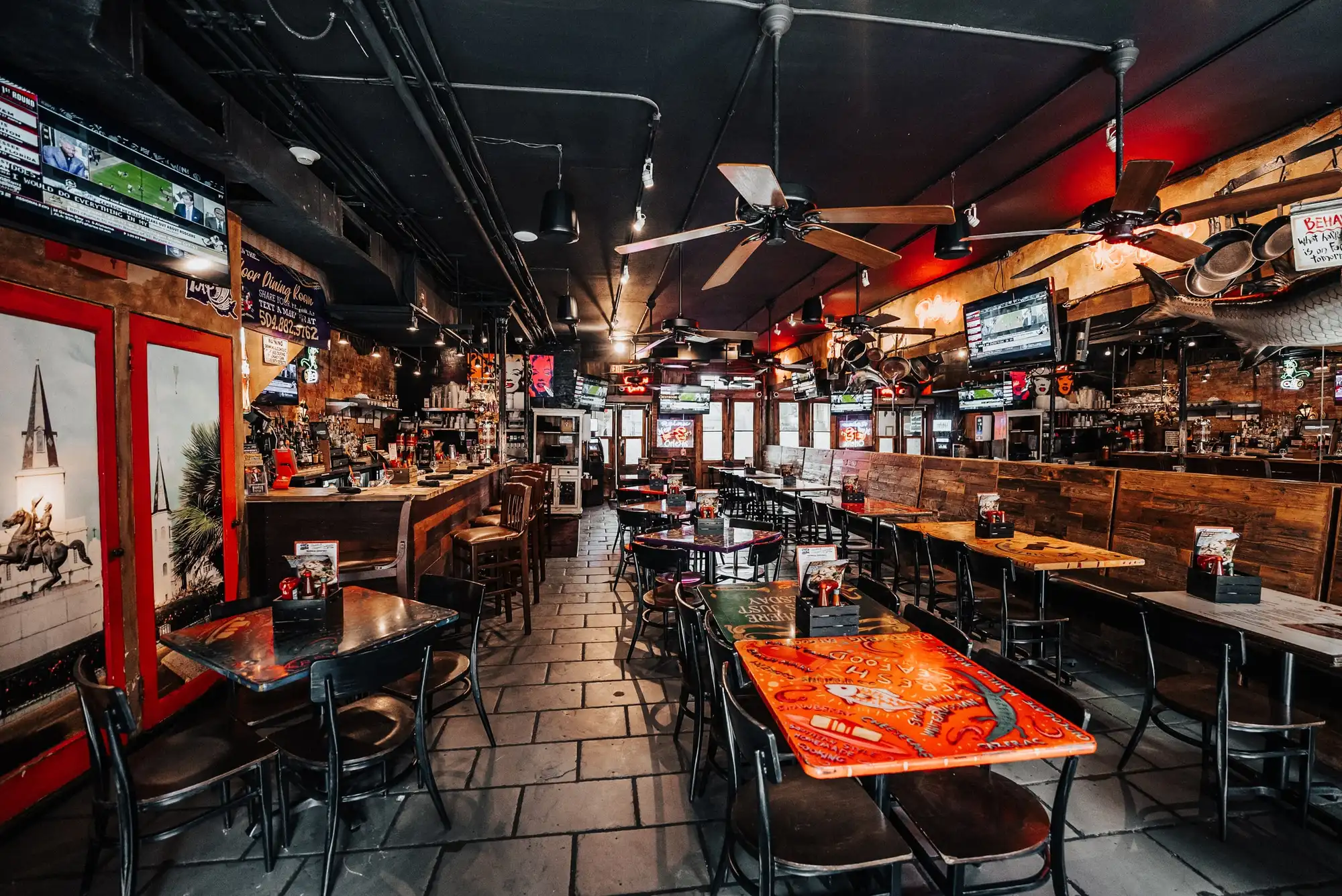 The long Main Dining Room at Olde Nola Cookery: painted tabletops, black chairs, ceiling fans and the bar running along the right